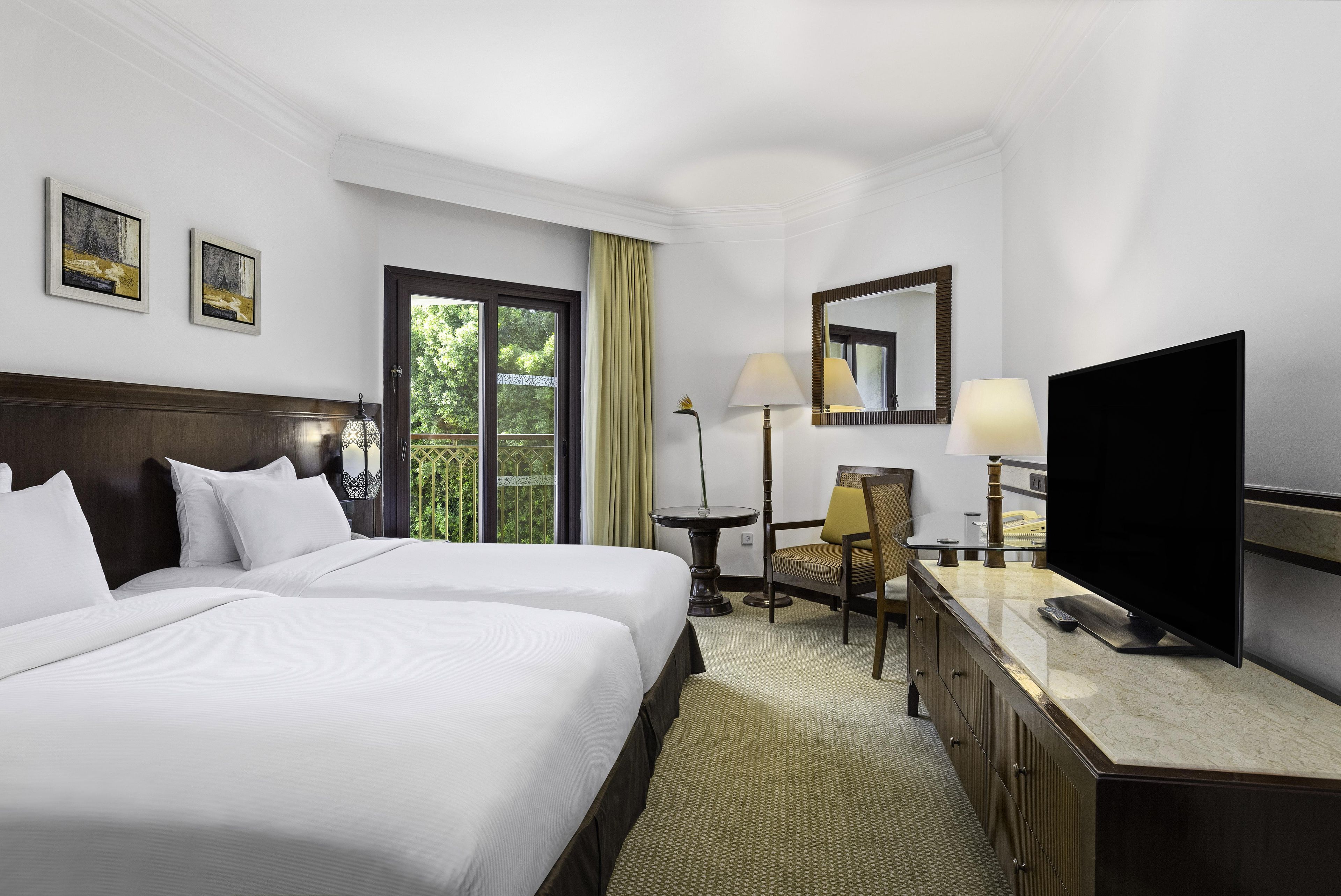 A hotel room with two beds, a desk with a TV, a chair, a lamp, a mirror, and artwork on the wall.