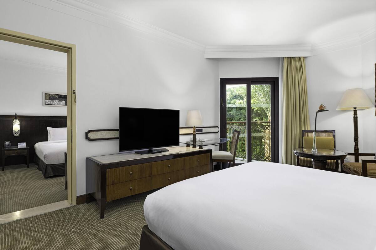 A hotel room with a bed, a television, a desk with a lamp, a chair, and a view of greenery through the window.