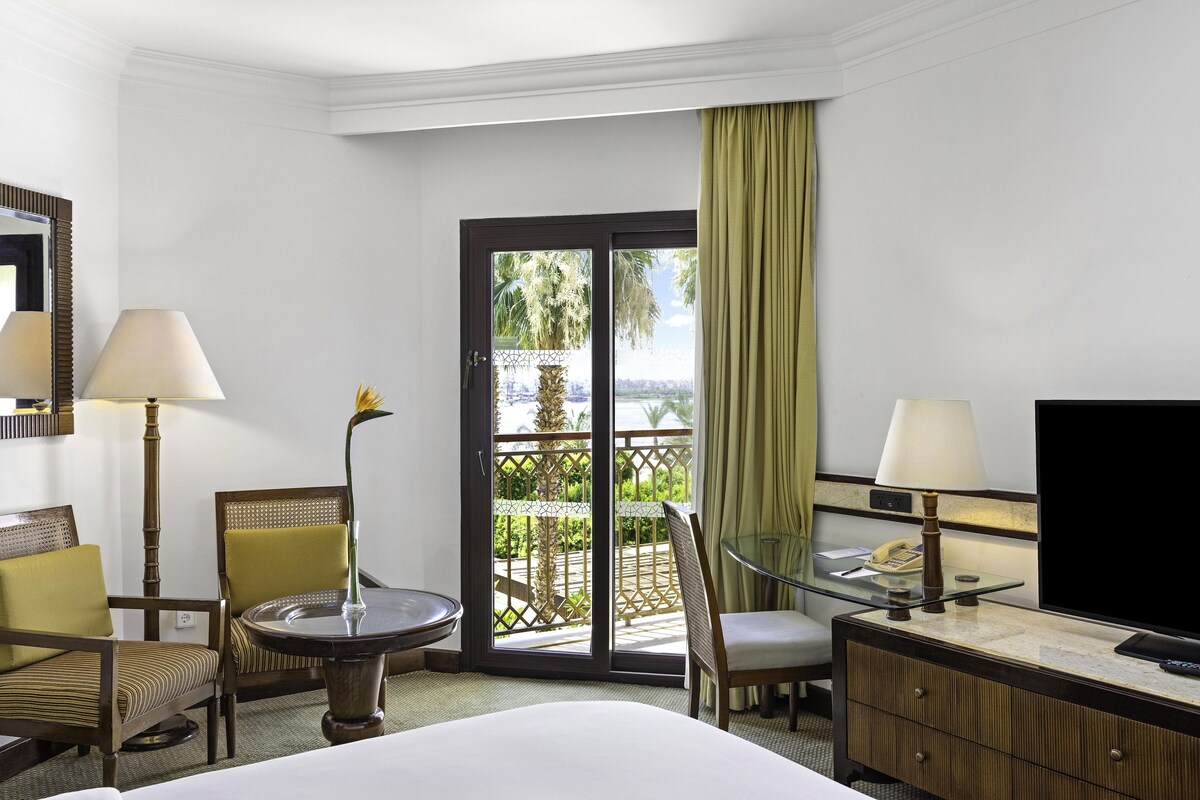 A hotel room with a bed, a desk with a lamp, a chair, a glass table, a television, and a sliding glass door leading to a balcony with a view.