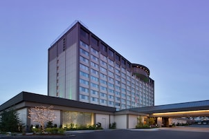 Un hotel de varios pisos con un diseño moderno, que cuenta con una entrada amplia con zona cubierta y un estacionamiento.