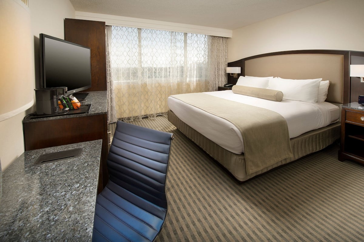 A hotel room with a bed, a desk with a flat-screen TV, a chair, and a window with curtains.