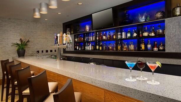 A bar area with a granite countertop, multiple bar stools, a variety of liquor bottles, and a flat-screen TV mounted above the bar. - Skybridge Seattle Airport Hotel (SeaTac)