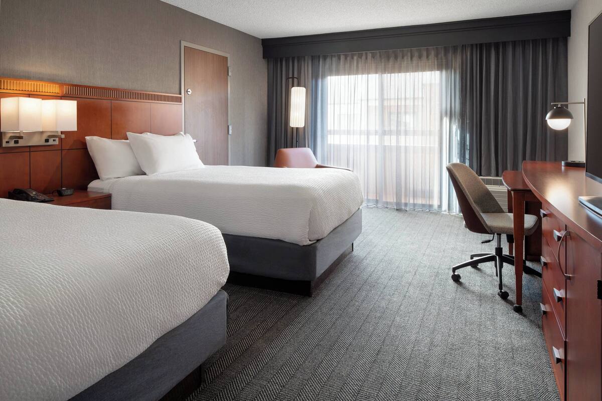 A hotel room with two beds, a desk, a chair, a lamp, and a large window with curtains.