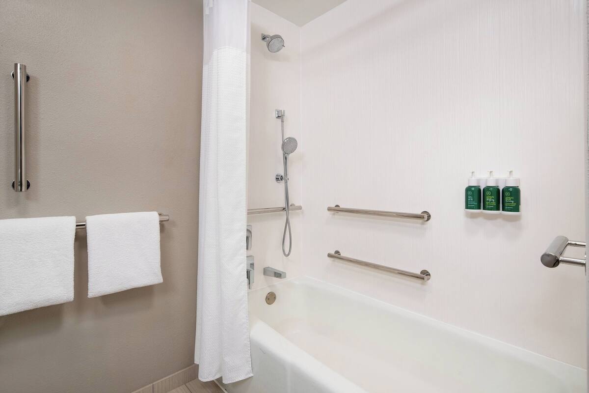 A modern bathroom with a bathtub, shower, and handrails. There are towels hanging on the wall and a shower curtain partially drawn.