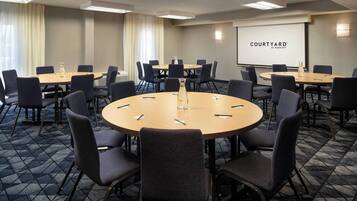 A conference room with multiple tables and chairs, a projector screen displaying 'COURTYARD BY MARIEOT', and a carpeted floor.