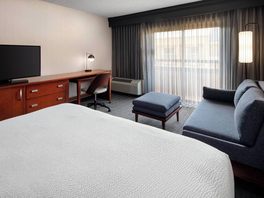 A hotel room with a large bed, a sofa, a desk with a chair, a television, and an air conditioning unit.