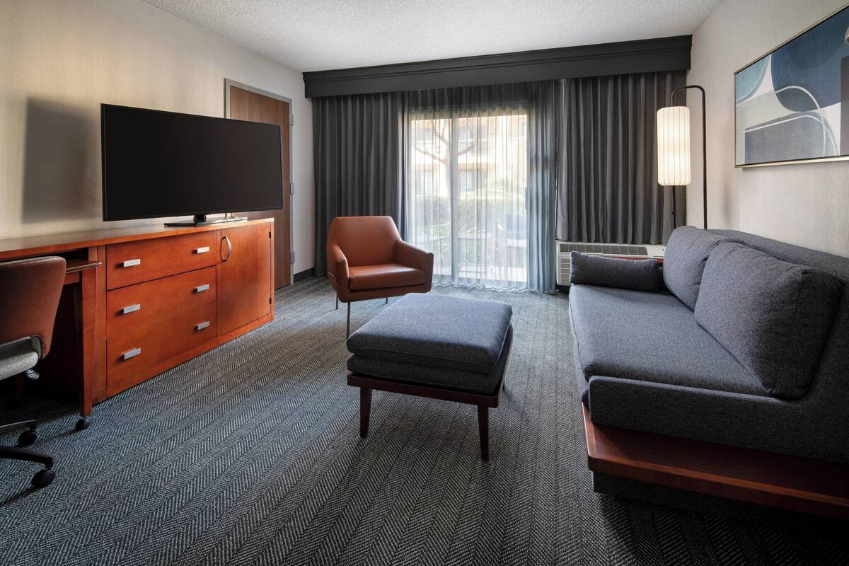 A hotel room with a flat-screen TV, a wooden desk with drawers, a leather armchair, a grey sofa, and a large window with curtains.