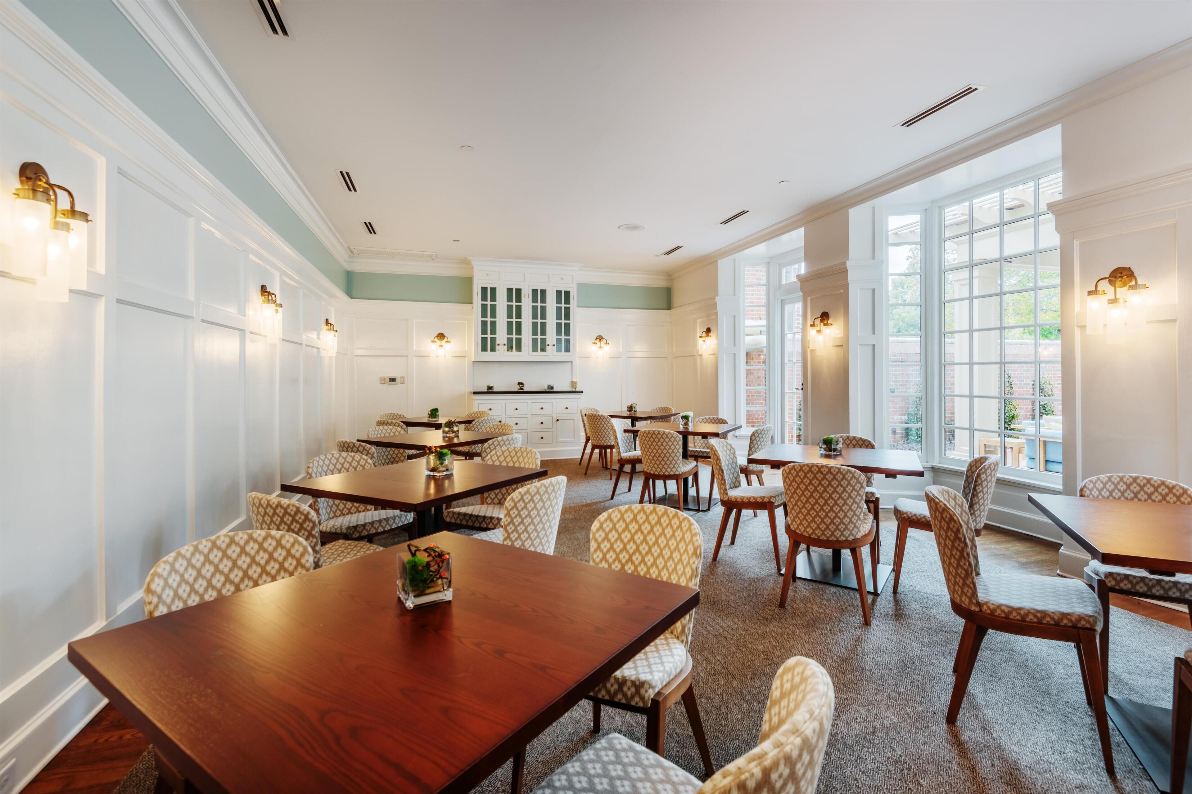 A spacious dining area with multiple tables and chairs, a kitchen in the background, and large windows allowing natural light.