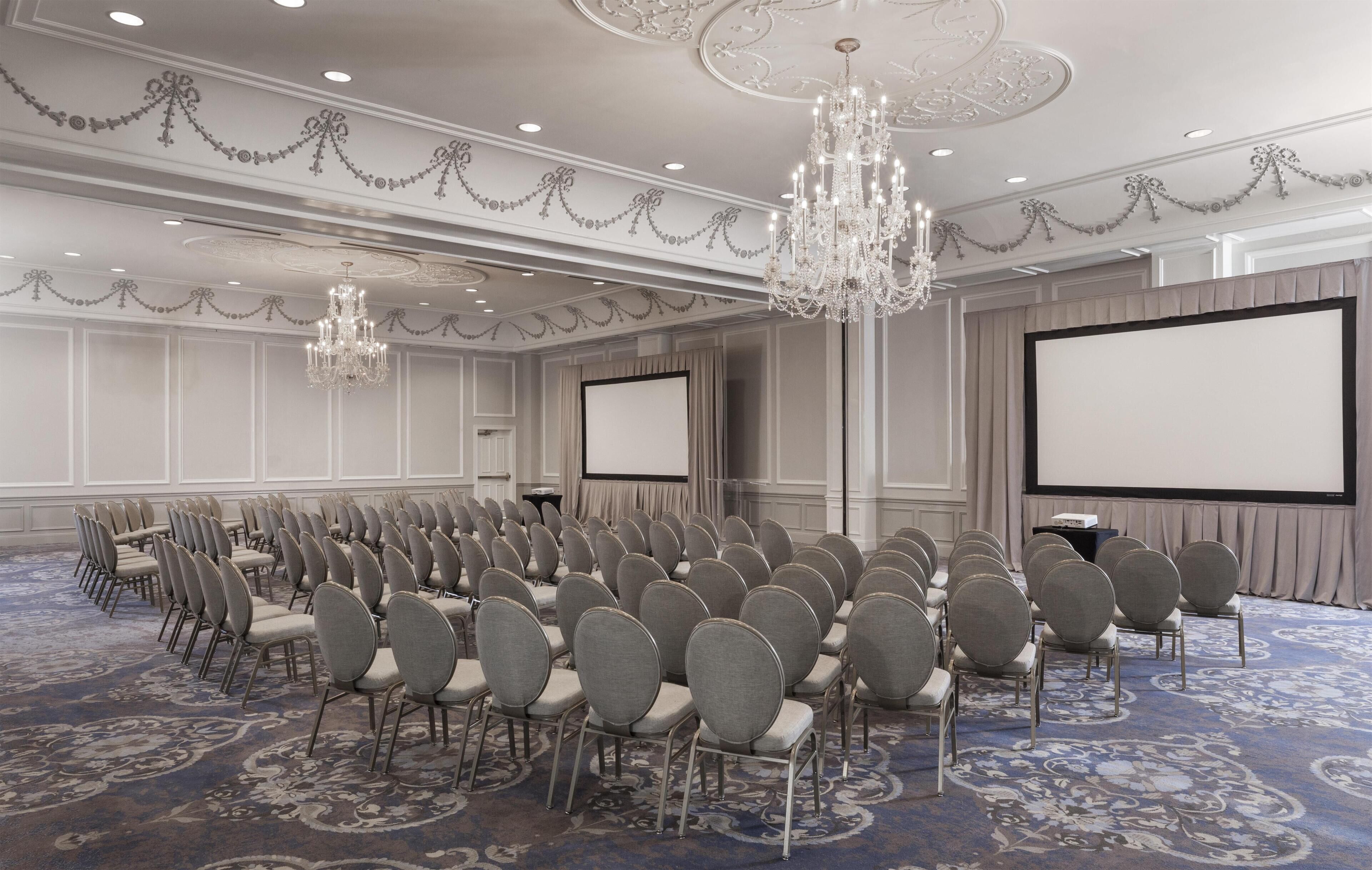 A conference room with rows of chairs, a large screen, and chandeliers.