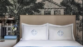 Una cama bien hecha con ropa blanca y almohadas decorativas, una mesita de noche con una lámpara y un jarrón de flores, y una pared con un mural detallado.