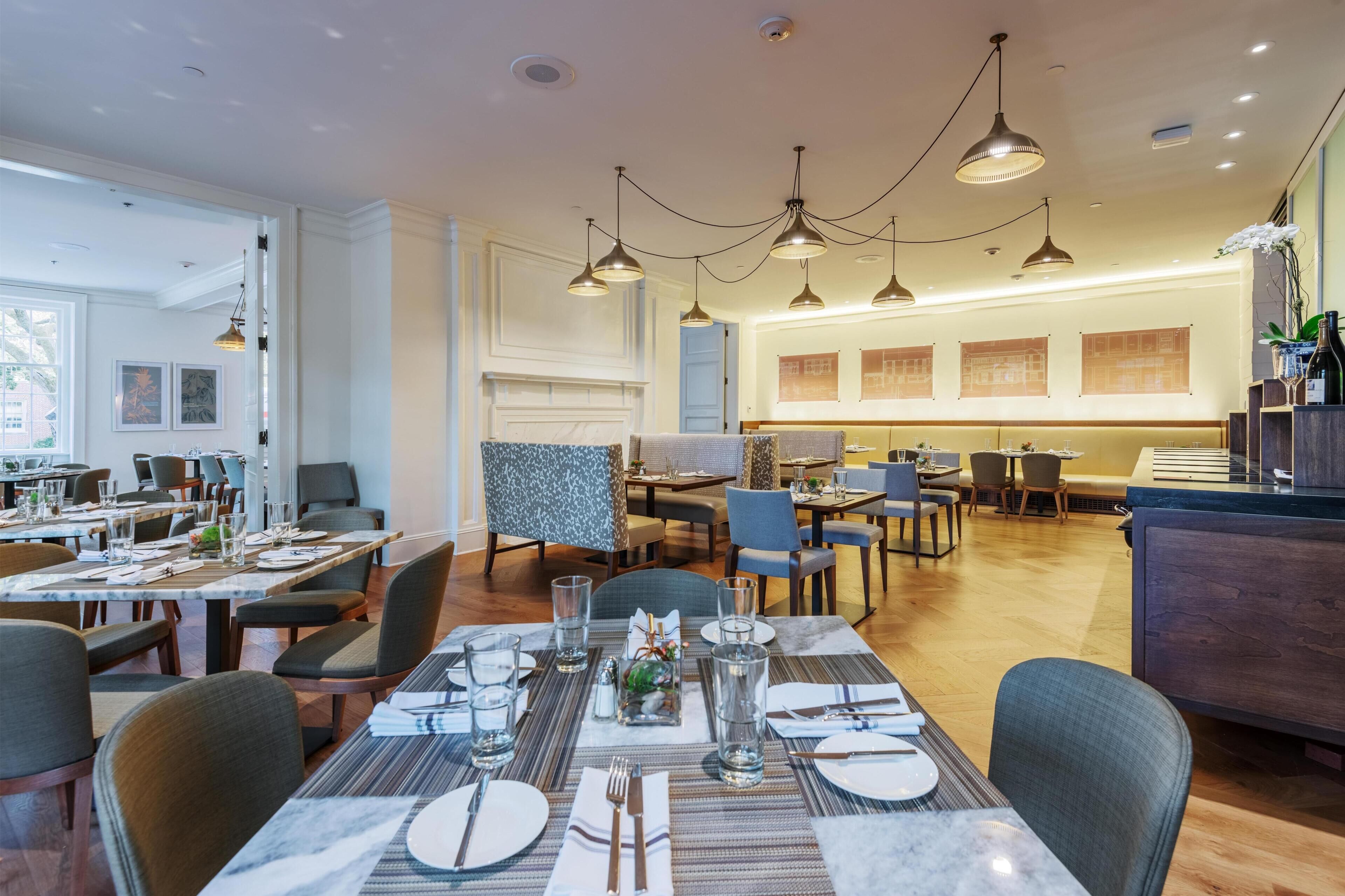 Un espace repas moderne, doté de plusieurs tables et chaises, d’un éclairage suspendu et offrant une vue sur la pièce adjacente.