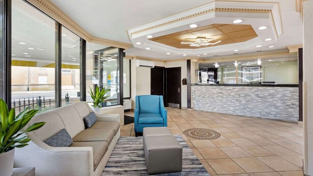 Eine Lobby mit einem beigen Sofa, einem blauen Sessel und einem grauen Hocker. Es gibt große Fenster, einen gemusterten Teppich und einen Fliesenboden.