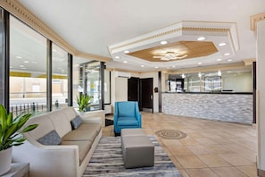 Eine Lobby mit einem beigen Sofa, einem blauen Sessel und einem grauen Hocker. Es gibt große Fenster, einen gemusterten Teppich und einen Fliesenboden.