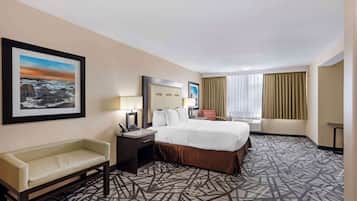 Habitación de hotel con una cama grande, un escritorio con silla, un sofá, una ventana con cortinas y un cuadro en la pared.