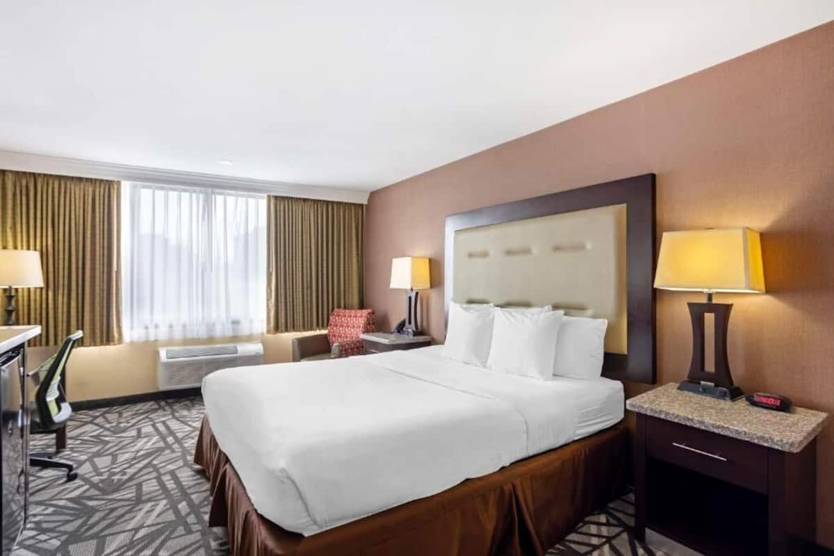 Habitación de hotel con una cama grande, dos mesitas de noche con lámparas, un escritorio con una silla, una silla roja y una ventana con cortinas.