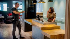 A modern reception desk with a wooden counter and a digital screen, a person standing in front of it, and another person walking by. - Hotel ibis Vannes (Vannes)