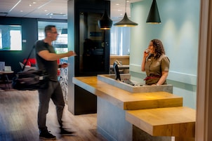 A modern reception desk with a wooden counter and a digital screen, a person standing in front of it, and another person walking by.