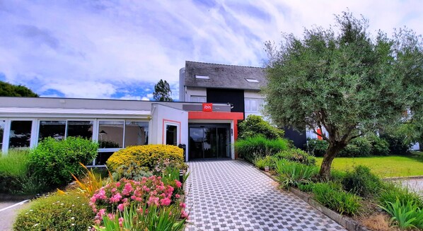 Un edificio moderno con fachada roja y blanca, entrada de vidrio y un sendero pavimentado que conduce a la entrada.