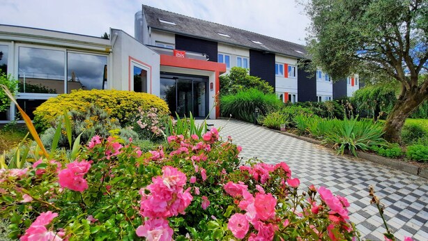 A modern building with large windows and a red and white facade. There is a well-maintained garden with pink flowers and a variety of greenery. - Hotel ibis Vannes (Vannes)