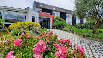 A modern building with large windows and a red and white facade. There is a well-maintained garden with pink flowers and a variety of greenery.
