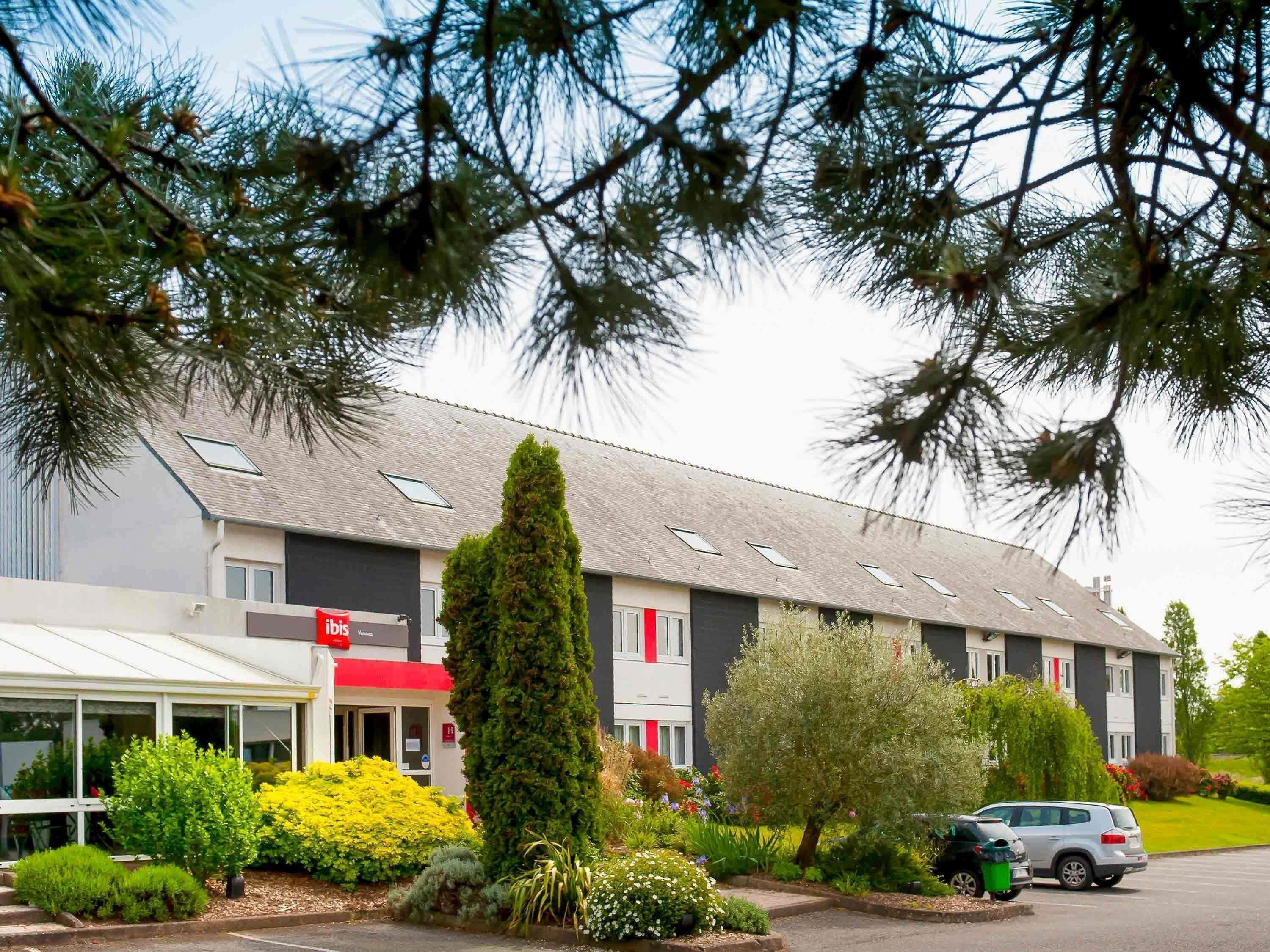 Un hotel de diseño moderno, con techo plano, numerosas ventanas y un jardín bien cuidado con arbustos variados y un alto árbol de coníferas.