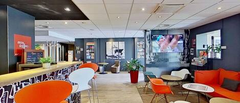 A modern hotel lobby with a reception desk, seating area, and a large TV screen.