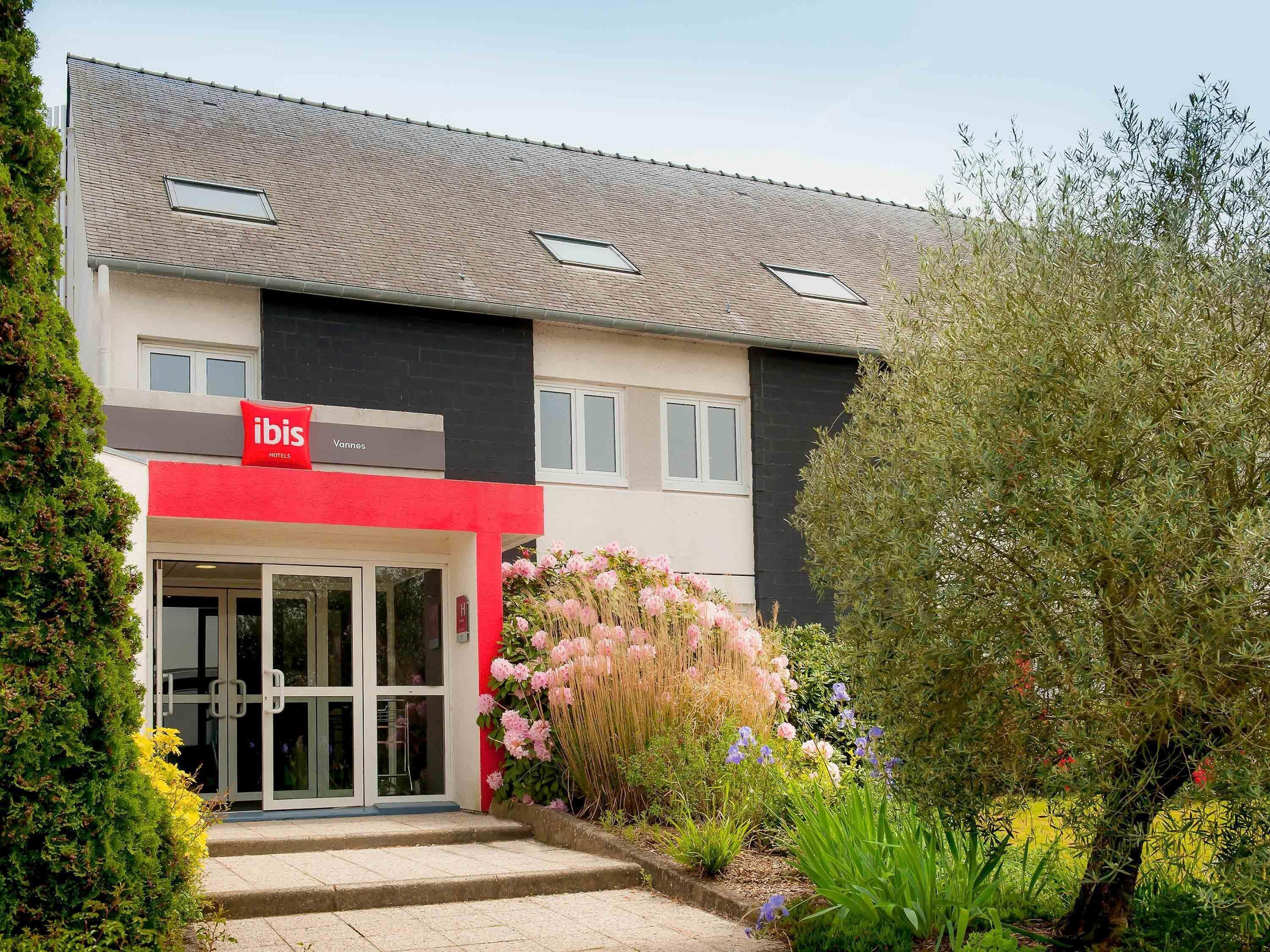 Un hôtel à la façade rouge et blanche, doté d’une entrée en verre et d’un jardin où poussent différentes plantes et fleurs.