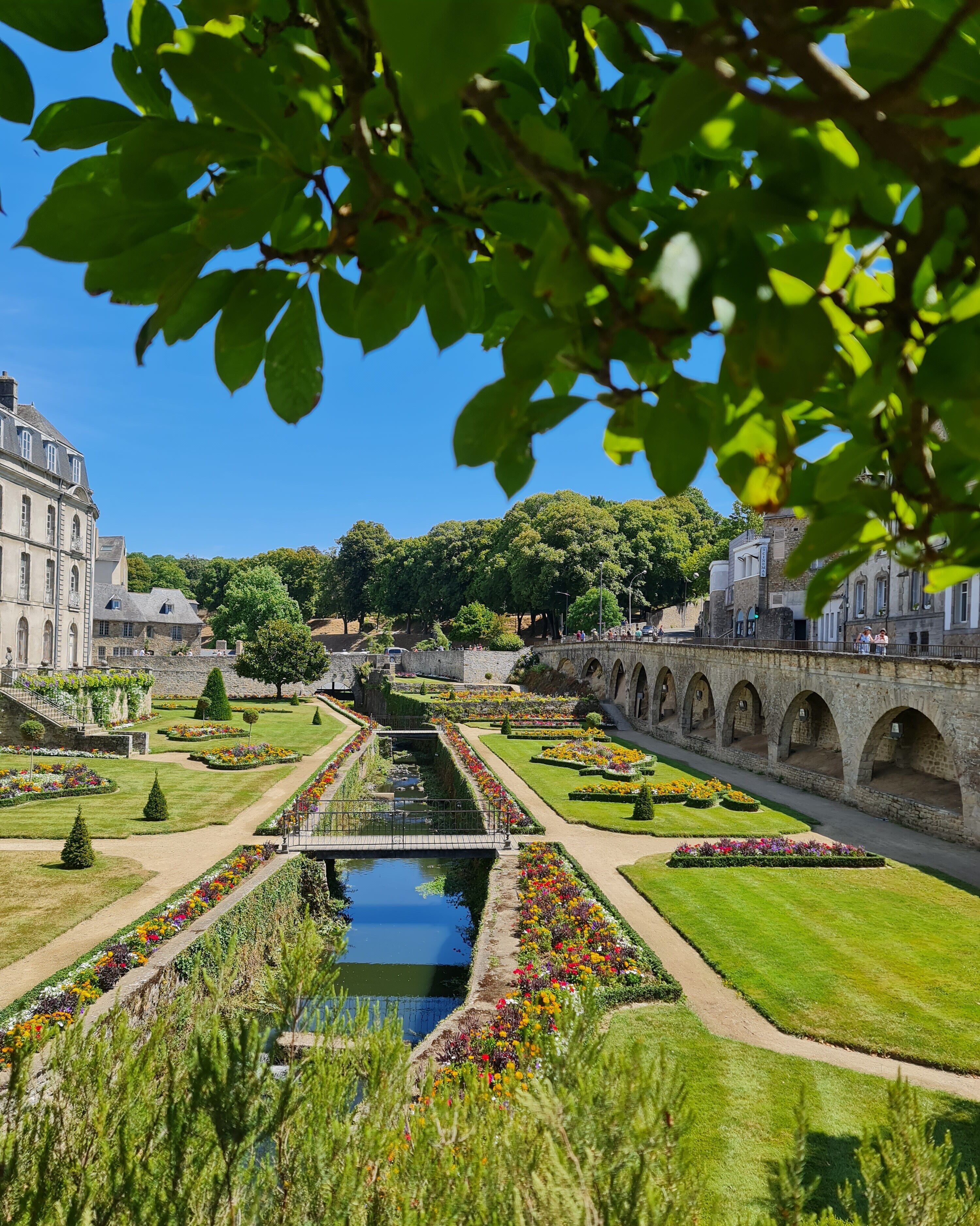 A formal garden with a central water feature, manicured lawns, and a stone bridge.