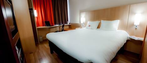 A hotel room with a large bed, a desk with a laptop, a red curtain, and a wooden floor.