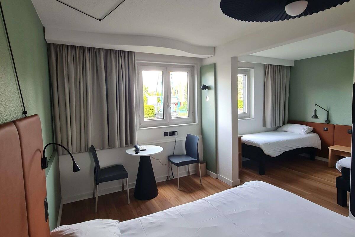 A hotel room with two beds, a desk, a chair, and two windows with curtains.