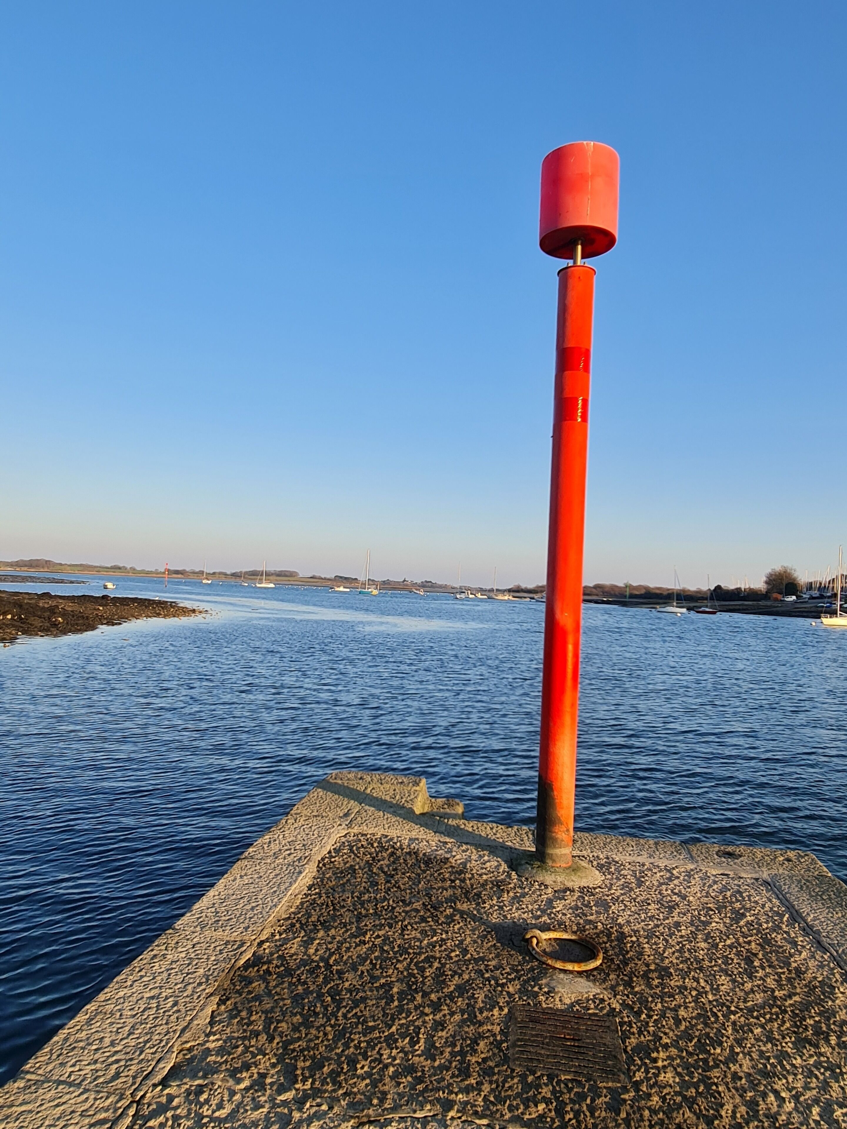 A pier with a red bollard and a view of the water and boats in the distance.