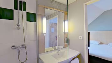 Ein Hotelbadezimmer mit Dusche, Waschbecken und einem durch eine Glastrennwand sichtbaren Bett.