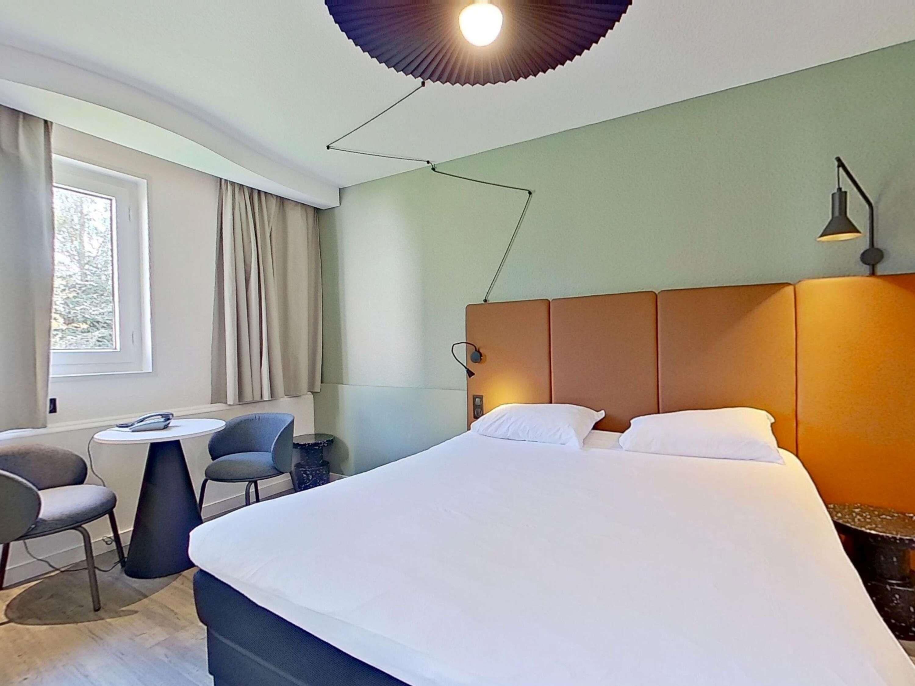 Ett modernt hotellrum med en stor säng, ett litet runt bord, två stolar, en lampa och ett fönster med gardiner.