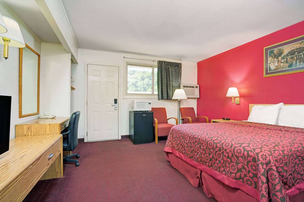 A hotel room with a bed, desk, chair, and a small refrigerator.