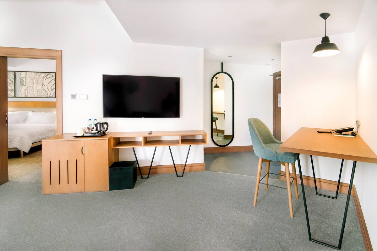 A modern hotel room with a bed, a desk with a chair, a TV, a mirror, and a lamp.