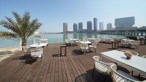 A deck with tables and chairs overlooking a body of water and a city skyline. - Le Méridien Abu Dhabi Resort (Abu Dhabi)