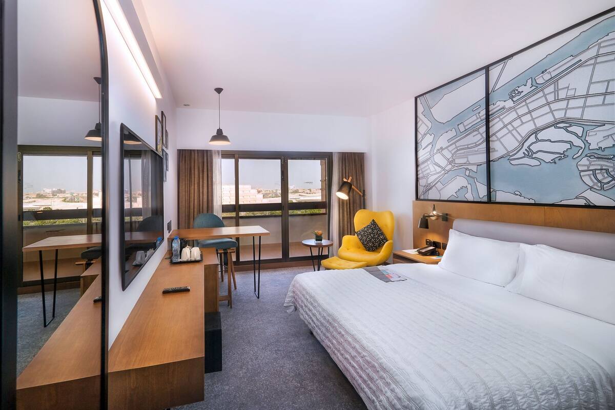 A hotel room with a large bed, a desk, a chair, and a view of the city through large windows.