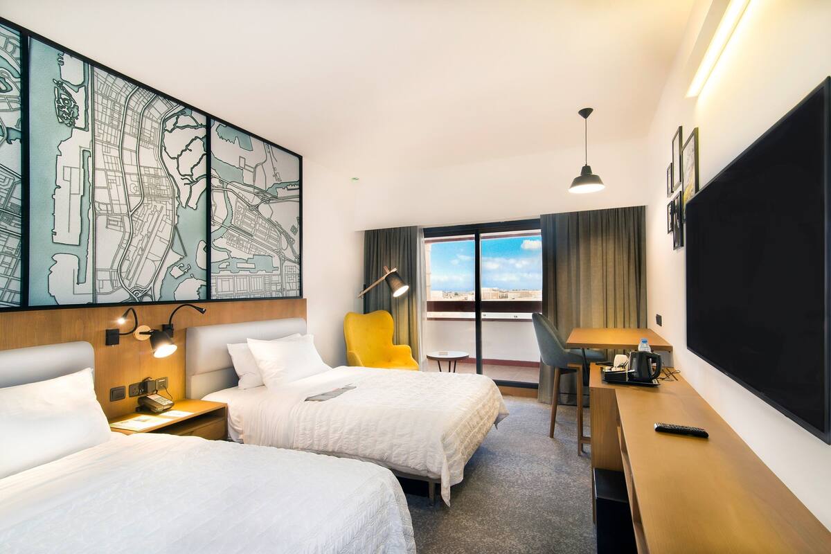 A hotel room with two beds, a desk, a chair, a large window, and a cityscape view.