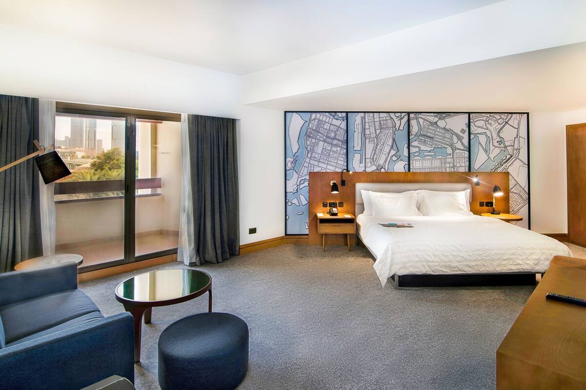 A hotel room with a bed, a desk, a chair, a small table, a nightstand, and a large window with curtains.