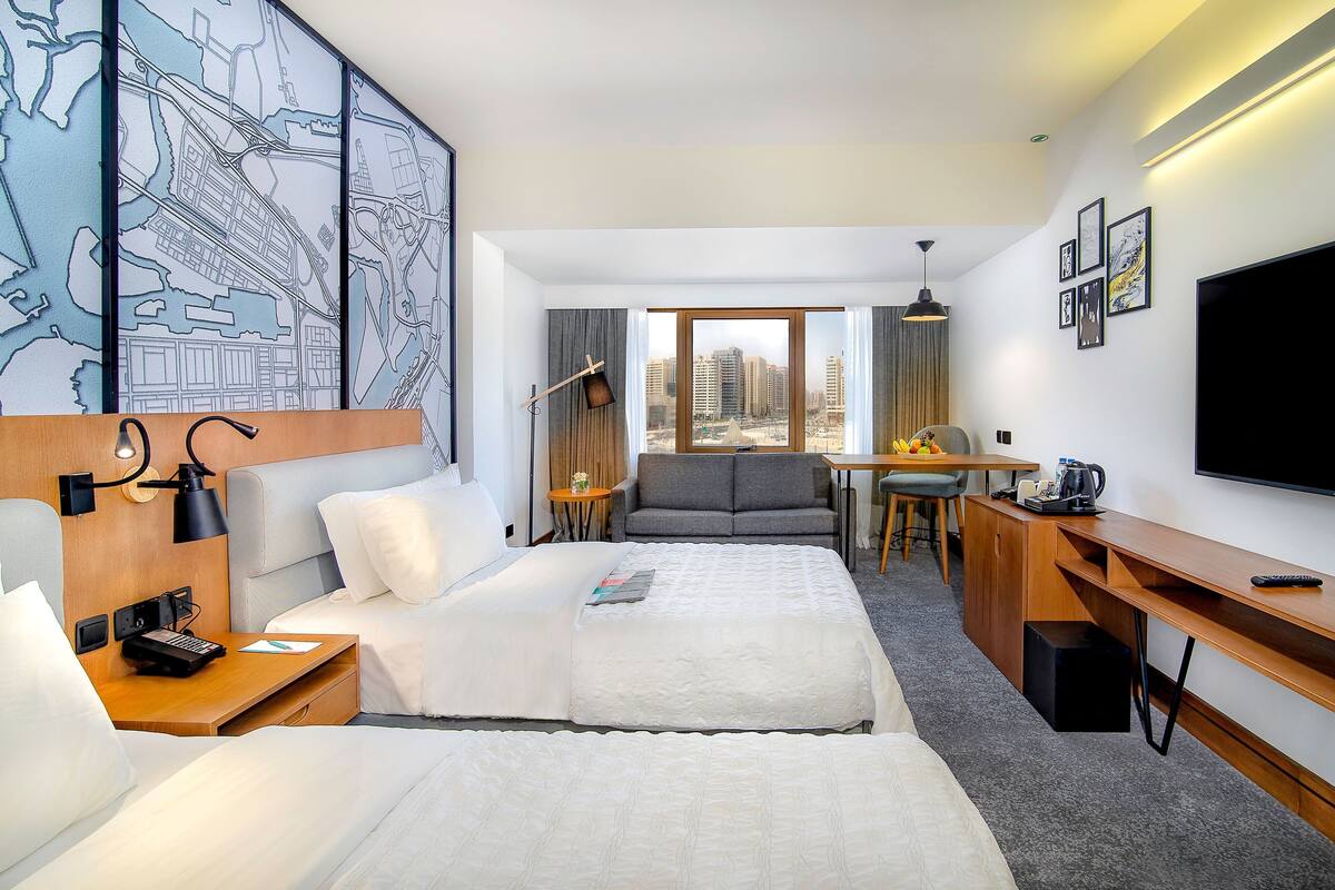 A hotel room with two beds, a desk, a chair, a TV, and a cityscape view.