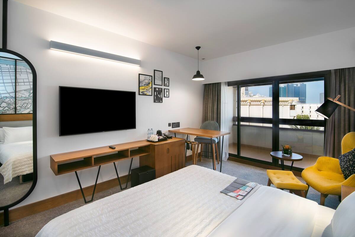 A hotel room with a bed, a desk, a chair, a TV, and a large window with a view of buildings.
