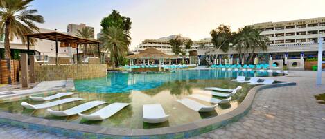 A swimming pool with lounge chairs and a covered seating area, surrounded by palm trees and a multi-story building in the background.