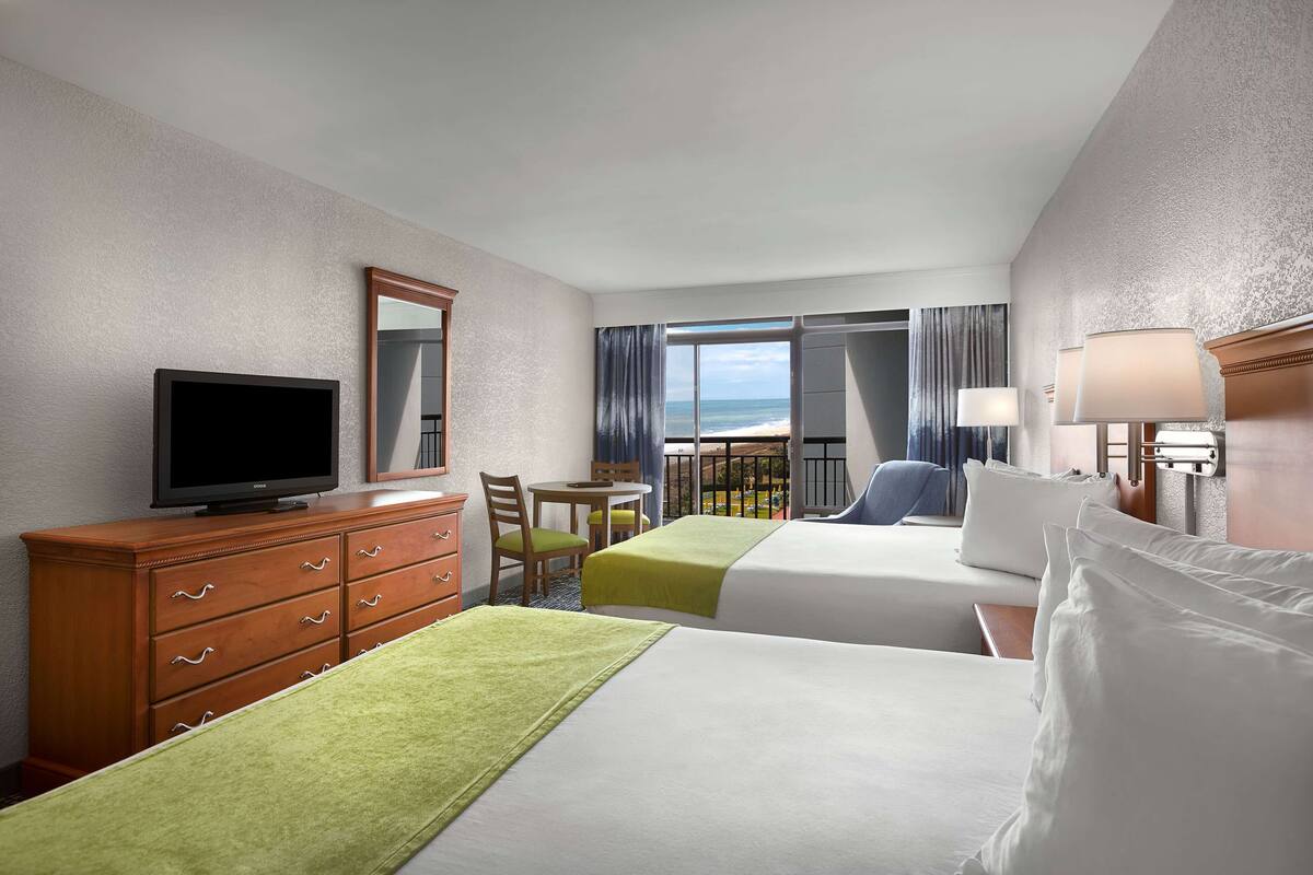 Une chambre d’hôtel avec deux lits, une télévision, une commode, une chaise, une petite table et un balcon donnant sur l  ’océan.