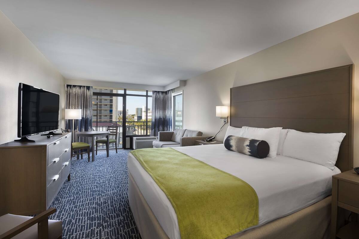 Une chambre d’hôtel avec un grand lit, un bureau avec une chaise, une télévision et une vue sur la ville grâce à de grandes fenêtres.