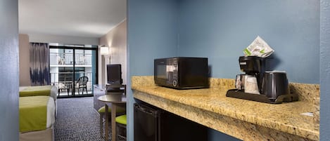 A compact hotel room with a kitchenette, a microwave, a coffee maker, and a refrigerator. There is a small dining table and a chair, and a view of the outside balcony.