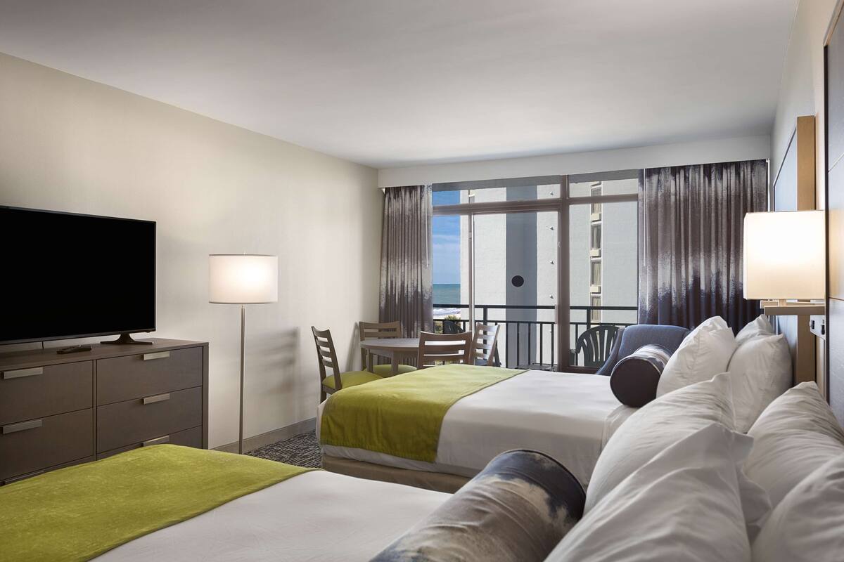 Une chambre d’hôtel avec deux lits, une télévision, un bureau et un balcon avec des rideaux.