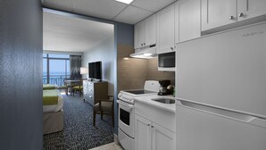 Quarto de hotel moderno com cozinha compacta, cama com manta verde, escrivaninha com cadeira e vista para o oceano.