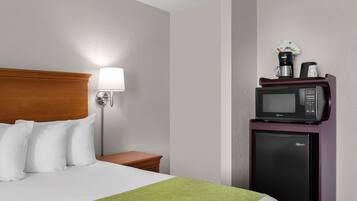 A hotel room with a bed, pillows, a nightstand, a lamp, a microwave, and a refrigerator.