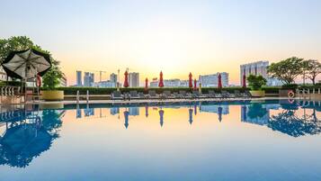 A swimming pool with lounge chairs and umbrellas, surrounded by greenery and high-rise buildings in the background.