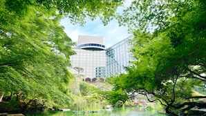 A modern building with a glass facade, surrounded by lush greenery and a calm water body.